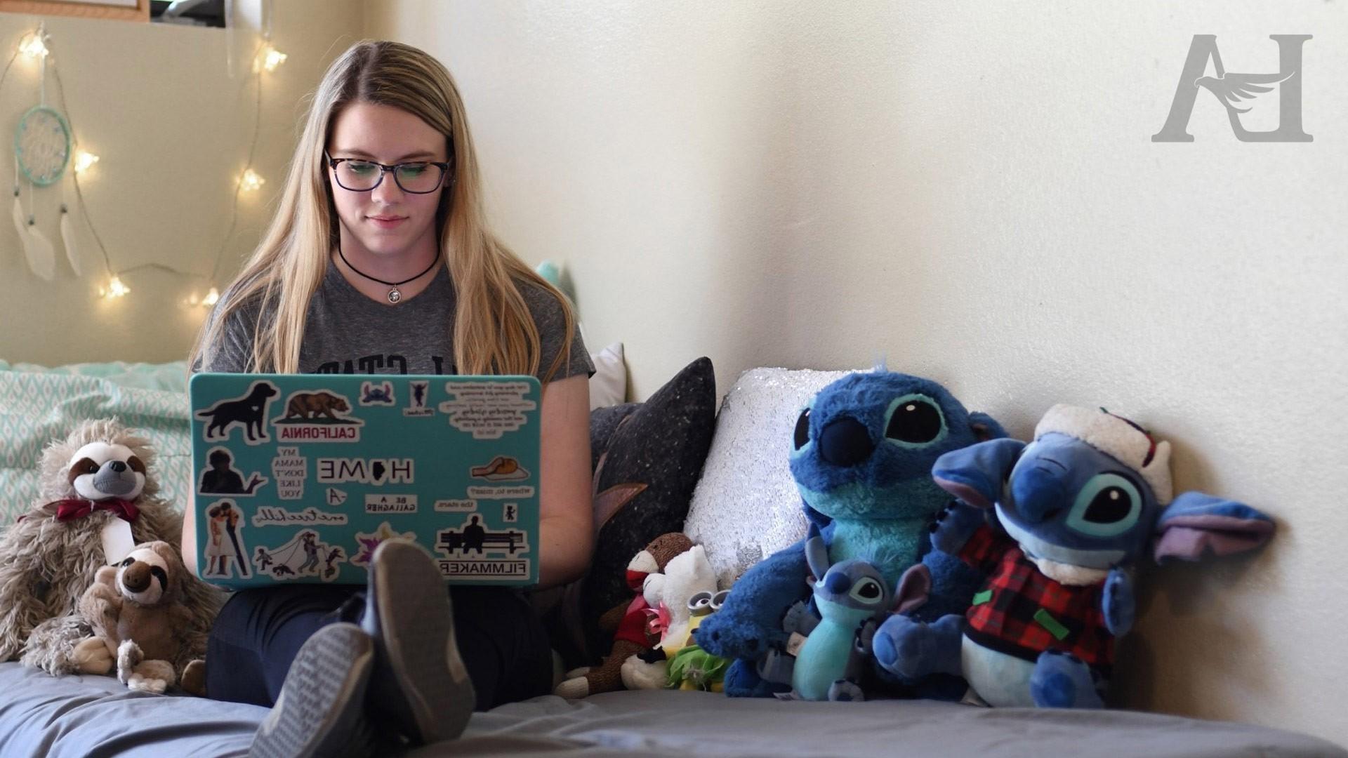 A student types on their laptop while sitting on their bed.